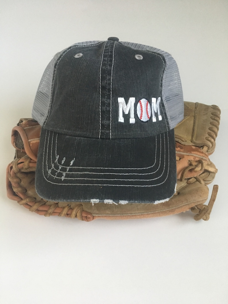 May include: A black and grey baseball cap with a white mesh back. The cap has the word "MOM" embroidered on the front with a red and white baseball graphic.