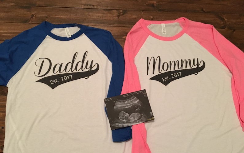 Puede incluir: Dos camisas blancas de manga larga con mangas azules y rosas. Las camisas tienen el texto "Daddy Est. 2017" y "Mommy Est. 2017" impreso en ellas. Una imagen de ultrasonido en blanco y negro est&aacute; en el centro de la foto.