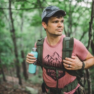 May include: A person wearing a red t-shirt with a mountain and trees graphic and the text "Explore More" is hiking in a forest. They are carrying a green backpack and a blue water bottle.