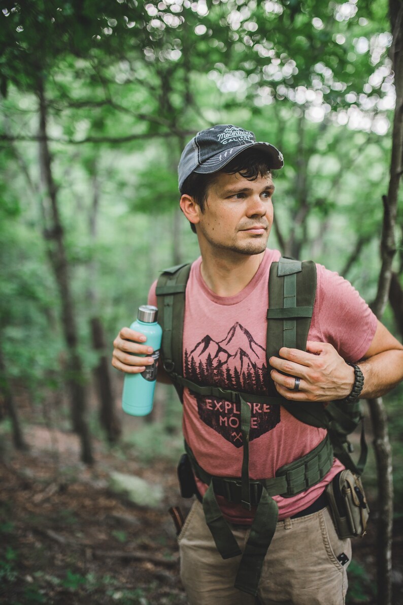 May include: A person wearing a red t-shirt with a mountain and trees graphic and the text "Explore More" is hiking in a forest. They are carrying a green backpack and a blue water bottle.