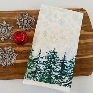 May include: A white kitchen towel with a winter forest scene printed on it. The scene features green pine trees with a snowy background. The towel is lying on a wooden cutting board with silver snowflake ornaments.
