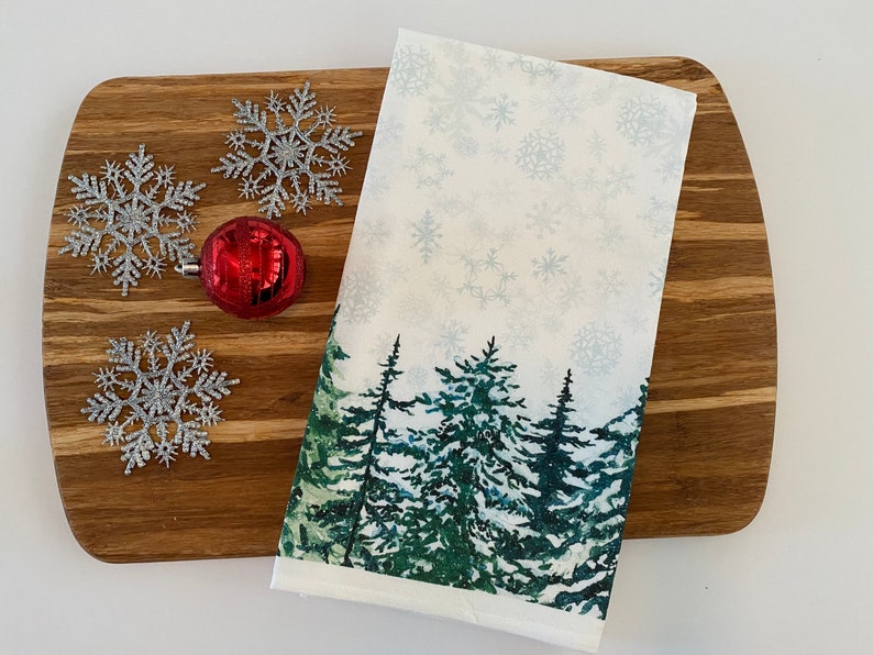 May include: A white kitchen towel with a winter forest scene printed on it. The scene features green pine trees with a snowy background. The towel is lying on a wooden cutting board with silver snowflake ornaments.