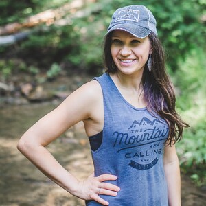 May include: A woman wearing a gray tank top with the text "The Mountains Are Calling And I Must Go" and a gray baseball cap with the text "Adventure" on it. She is standing in a forest setting.