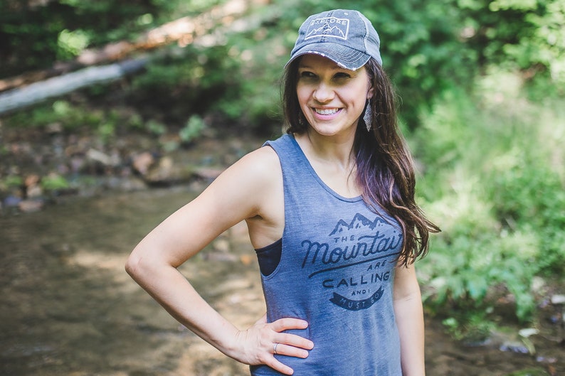 May include: A woman wearing a gray tank top with the text "The Mountains Are Calling And I Must Go" and a gray baseball cap with the text "Adventure" on it. She is standing in a forest setting.