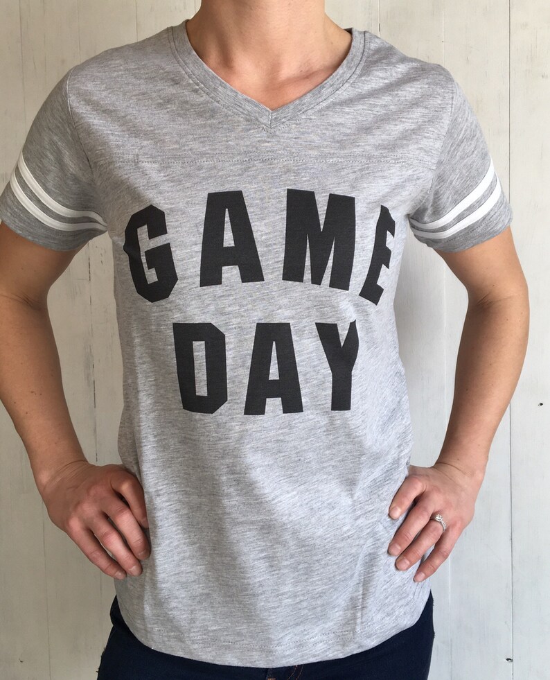 May include: A gray v-neck t-shirt with white stripes on the sleeves. The shirt has the text "GAME DAY" printed in black on the front.