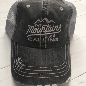 Puede incluir: Una gorra de camionero negra y gris desgastada con una parte trasera de malla blanca. La gorra tiene bordado blanco que dice "The Mountains Are Calling".