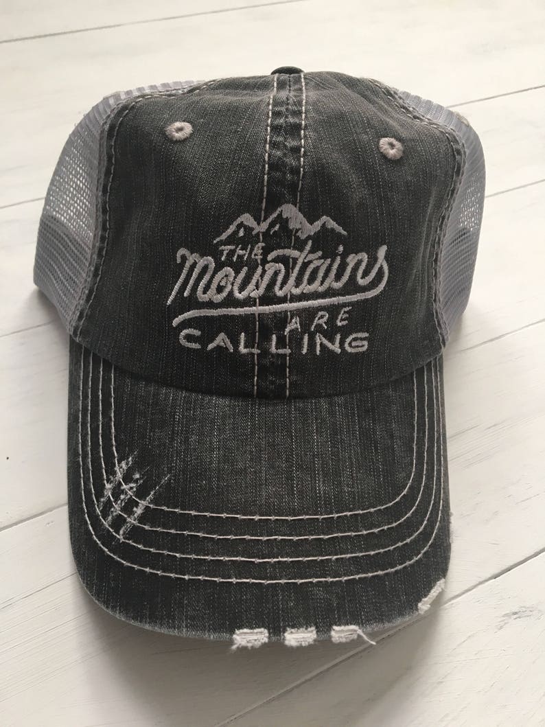 Puede incluir: Una gorra de camionero negra y gris desgastada con una parte trasera de malla blanca. La gorra tiene bordado blanco que dice "The Mountains Are Calling".