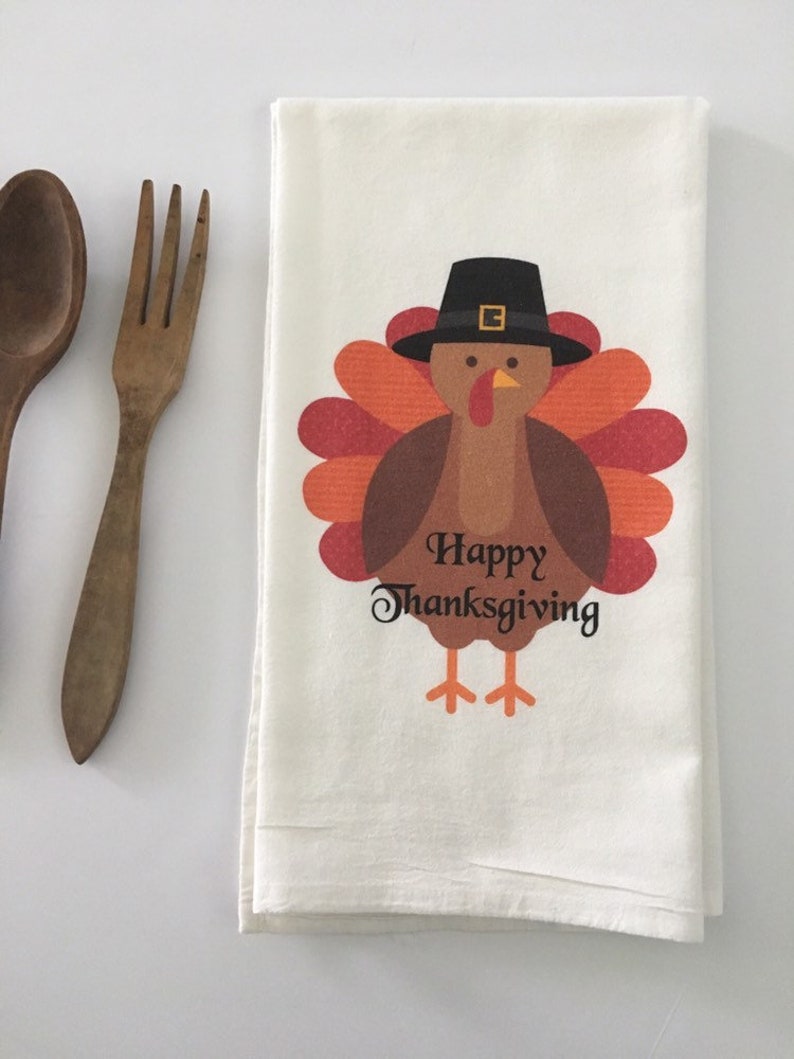 Puede incluir: Una toalla de cocina blanca con un dibujo de un pavo que lleva un sombrero de peregrino. El pavo es marr&oacute;n con plumas naranjas y rojas. El texto "Happy Thanksgiving" est&aacute; impreso debajo del pavo.