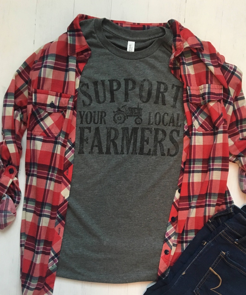 May include: A grey t-shirt with the text "Support Your Local Farmers" printed in black. The shirt is layered over a red and black checked flannel shirt and blue jeans.