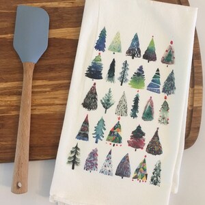 K&ouml;nnte beinhalten: Ein wei&szlig;es Geschirrtuch mit einem farbenfrohen Aquarelldesign aus verschiedenen Weihnachtsb&auml;umen. Die B&auml;ume sind in Gr&uuml;n-, Blau-, Rot- und Gelbt&ouml;nen gemalt.