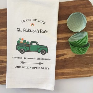 Könnte beinhalten: Weißes Küchentuch mit einem grünen Lastwagen, der Kürbisse, Kleeblätter und Regenbögen transportiert. Der Text lautet "Loads of Luck St. Patrick's Fair Clovers - Rainbows - Leprechauns One Mile - Open Daily".
