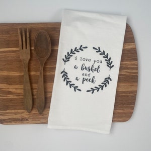 May include: A white kitchen towel with the text "I love you a bushel and a peck" in black script, surrounded by a wreath design. The towel is on a wooden cutting board with a wooden fork and spoon.