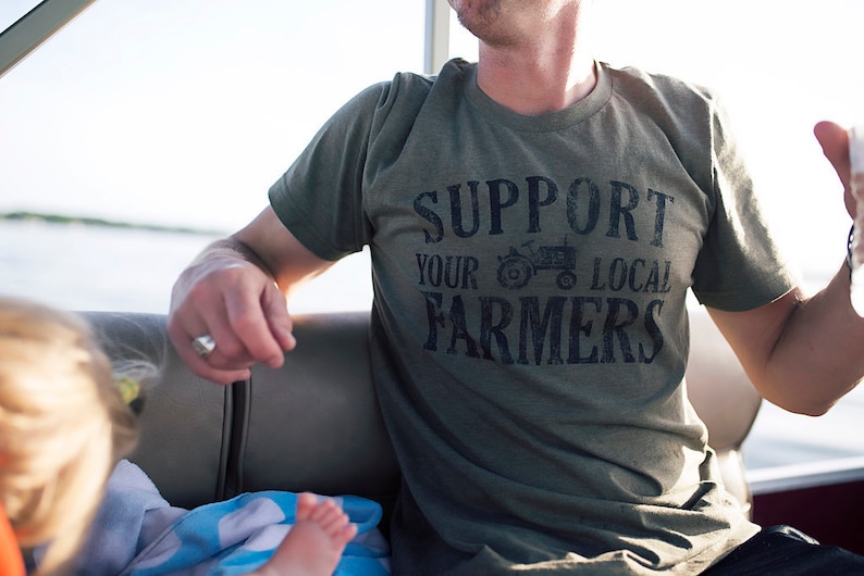 May include: A man wearing a green t-shirt with the text "Support Your Local Farmers" and a small tractor graphic.