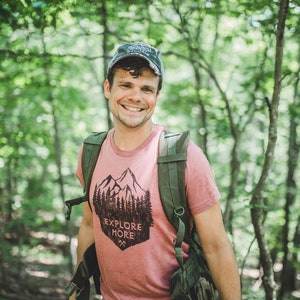 May include: A man wearing a pink t-shirt with a mountain and trees graphic and the text "Explore More". He is wearing a green backpack and a blue baseball cap. He is standing in a forest.