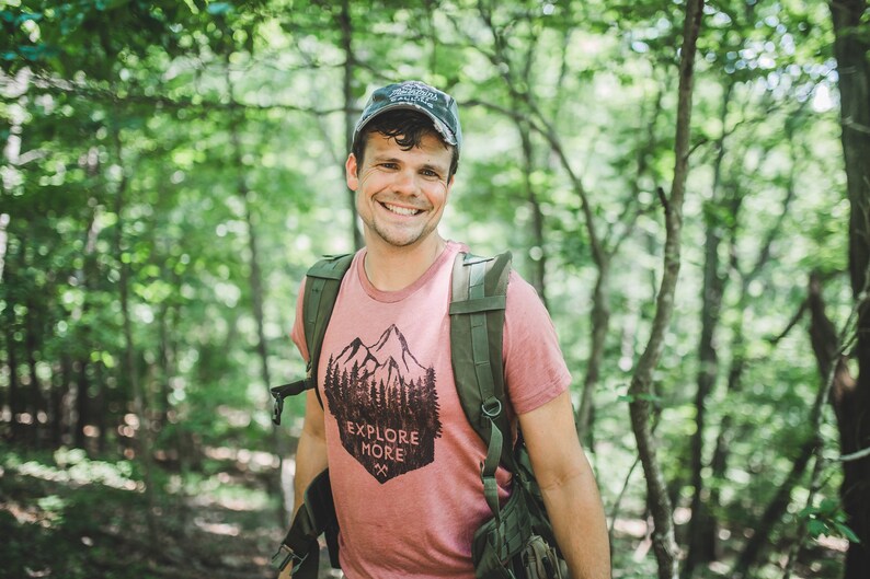 May include: A man wearing a pink t-shirt with a mountain and trees graphic and the text "Explore More". He is wearing a green backpack and a blue baseball cap. He is standing in a forest.