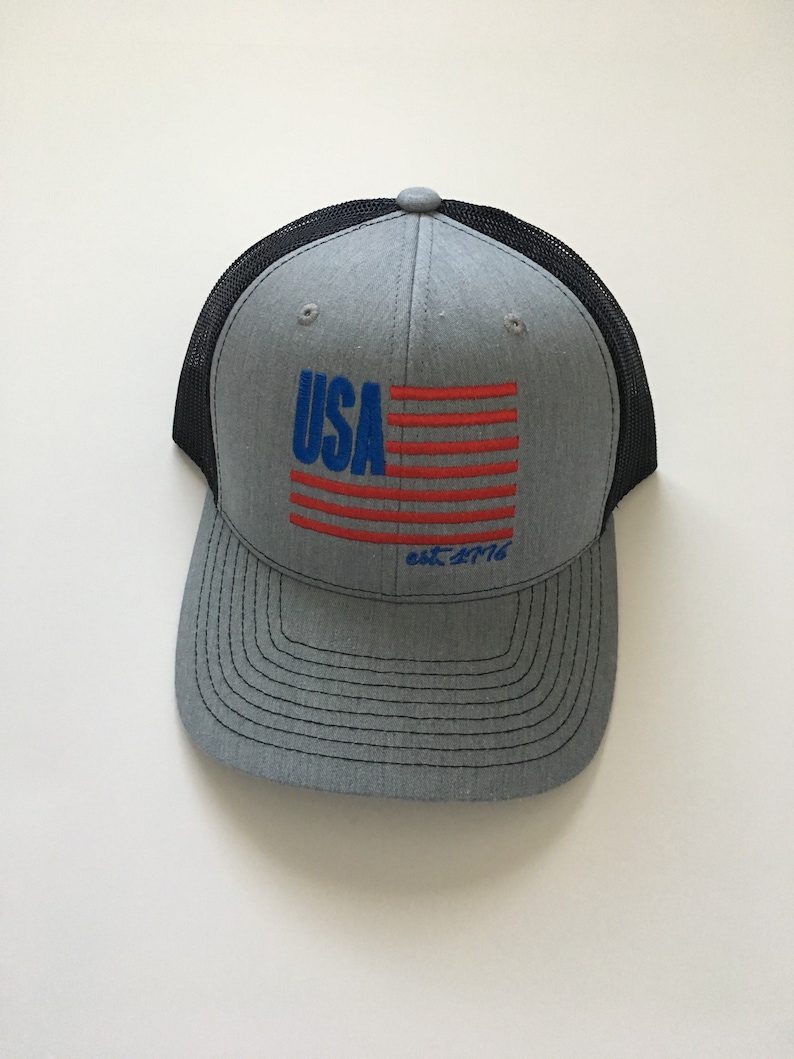 May include: A grey and black trucker hat with a red, white and blue American flag design and the text "USA est. 1776".