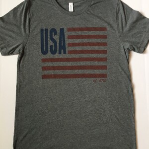 Pode incluir: Uma camiseta cinza mescla com um design de bandeira americana azul e vermelha. O texto "USA est. 1776" est&aacute; impresso na camiseta.
