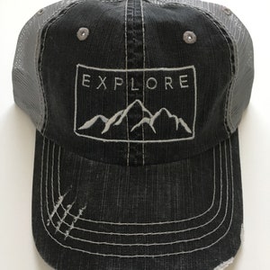May include: A black and gray distressed trucker hat with a white embroidered mountain range and the word "EXPLORE" in a white rectangle.
