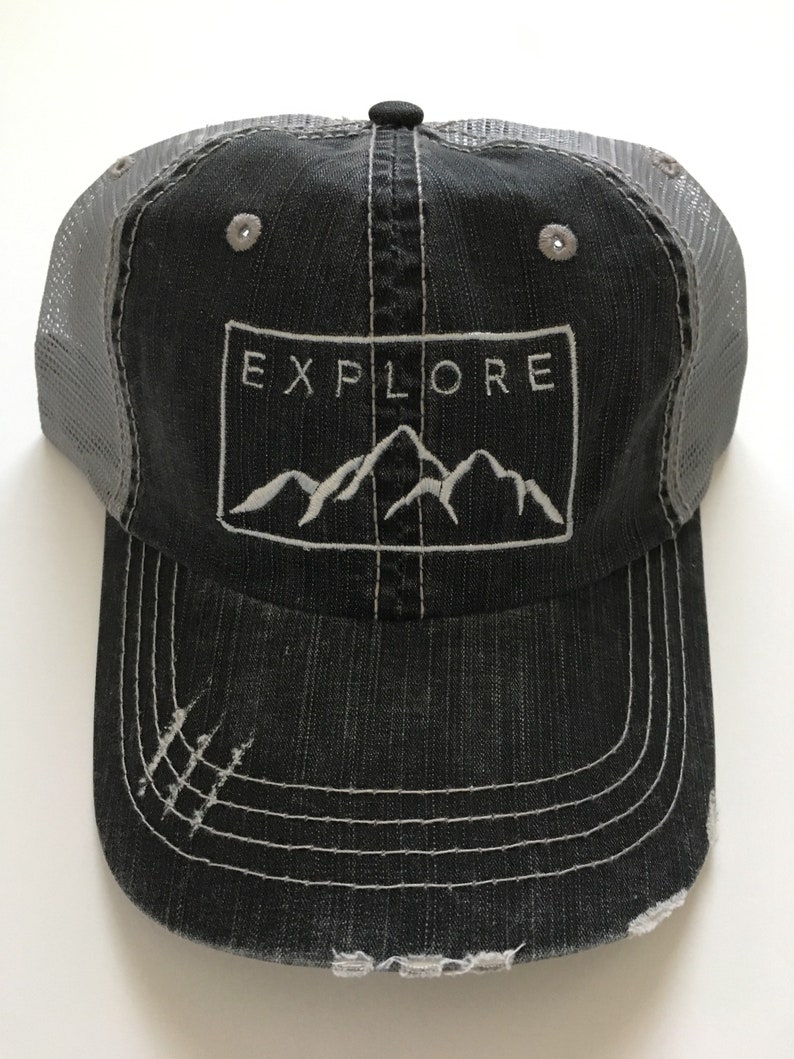 May include: A black and gray distressed trucker hat with a white embroidered mountain range and the word "EXPLORE" in a white rectangle.