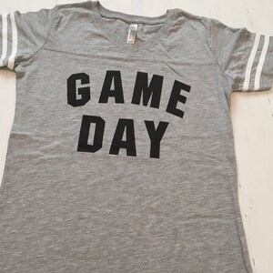 May include: Gray V-neck t-shirt with white stripes on the sleeves and the words "GAME DAY" printed in black.
