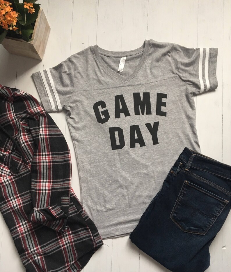 May include: Gray V-neck t-shirt with white stripes on the sleeves and the text "GAME DAY" printed in black.