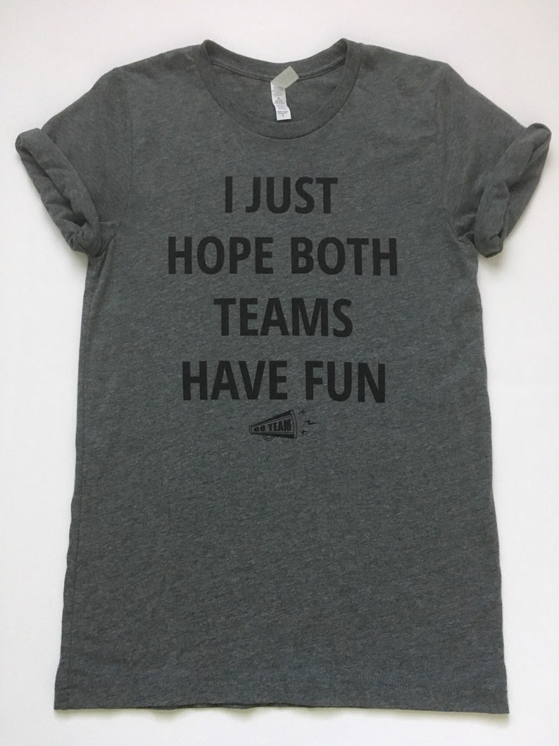 May include: A heather gray t-shirt with the text "I JUST HOPE BOTH TEAMS HAVE FUN" printed in black. A small black and white graphic of a megaphone with the text "GO TEAM" is below the text.