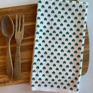 May include: A white kitchen towel with a green shamrock pattern. The towel is folded and lying on a wooden cutting board.