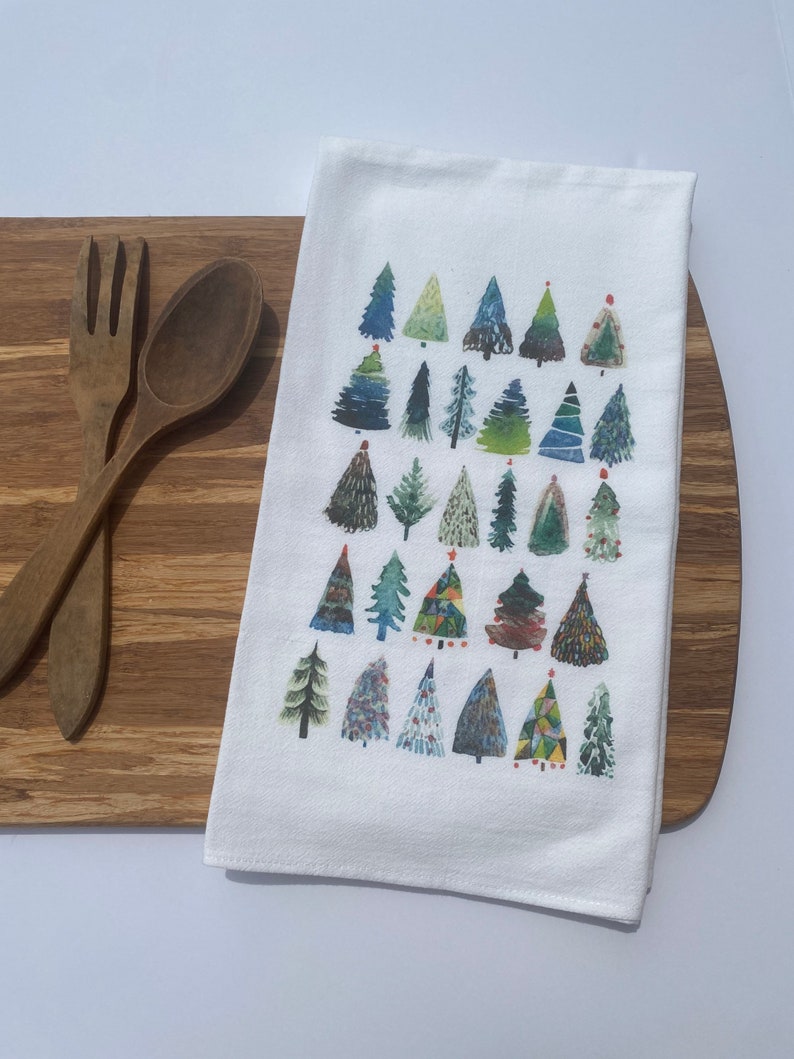 K&ouml;nnte beinhalten: Ein wei&szlig;es K&uuml;chentuch mit einem farbenfrohen Aquarelldesign aus verschiedenen Weihnachtsb&auml;umen. Die B&auml;ume sind in Gr&uuml;n-, Blau- und Rott&ouml;nen gemalt.