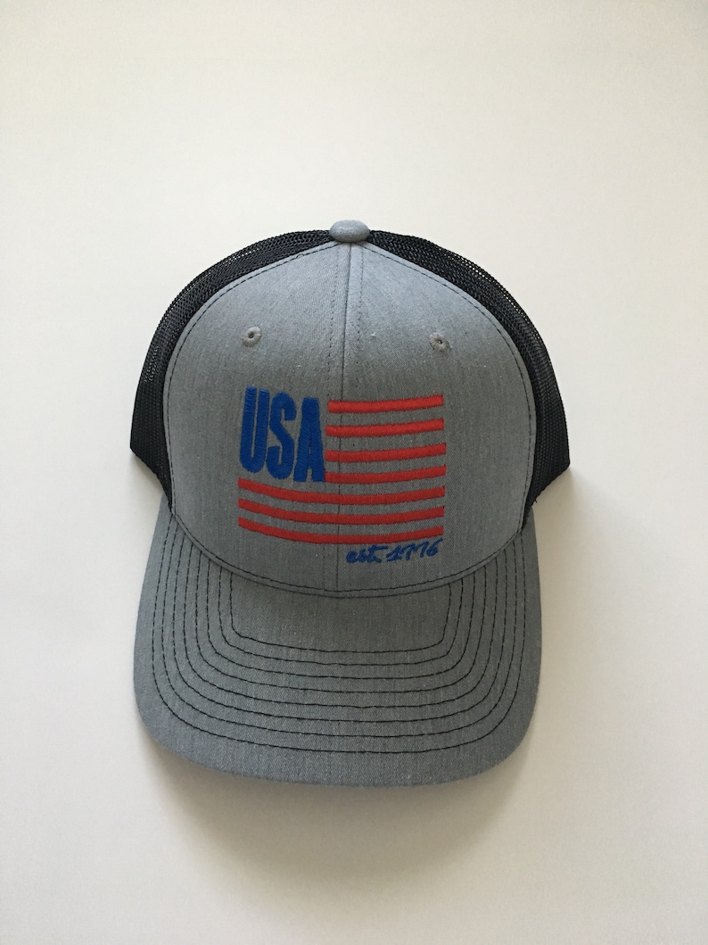 May include: A grey and black trucker hat with a red, white and blue American flag design and the text "USA est. 1776".