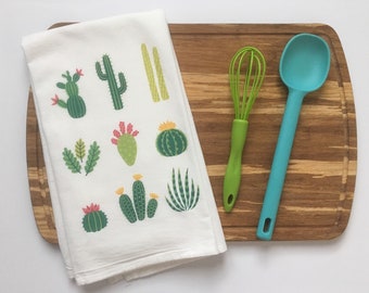 May include: A white kitchen towel with a cactus print design. The towel is folded and placed on a wooden cutting board with a green whisk and a blue silicone spoon.