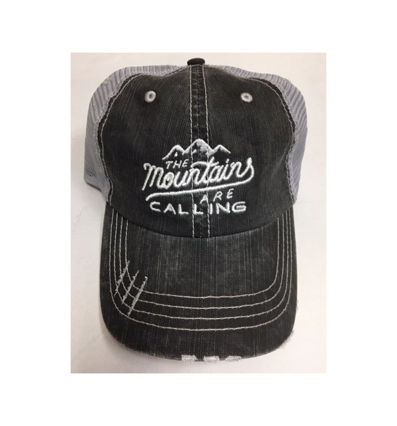 Puede incluir: Una gorra de camionero negra y gris con un dise&ntilde;o bordado en blanco que dice "The Mountains Are Calling".