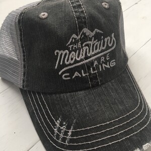 Puede incluir: Una gorra de camionero negra y gris desgastada con texto bordado en blanco que dice "The Mountains Are Calling".