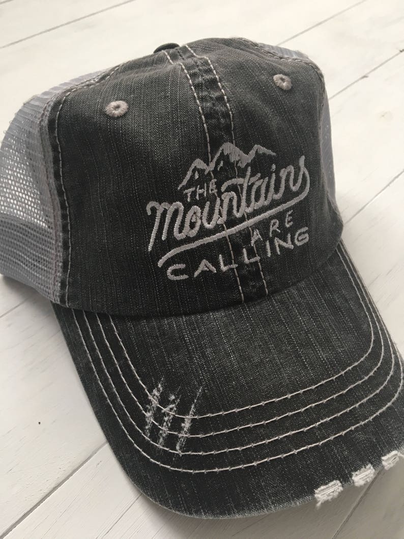 Puede incluir: Una gorra de camionero negra y gris desgastada con texto bordado en blanco que dice "The Mountains Are Calling".