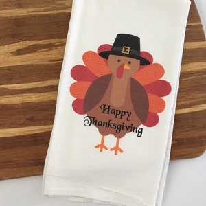 Puede incluir: Toalla de cocina blanca con un dibujo de un pavo de dibujos animados que lleva un sombrero de peregrino. El pavo tiene plumas marrones y una papada roja y naranja. El texto "Happy Thanksgiving" est&aacute; impreso debajo del pavo.