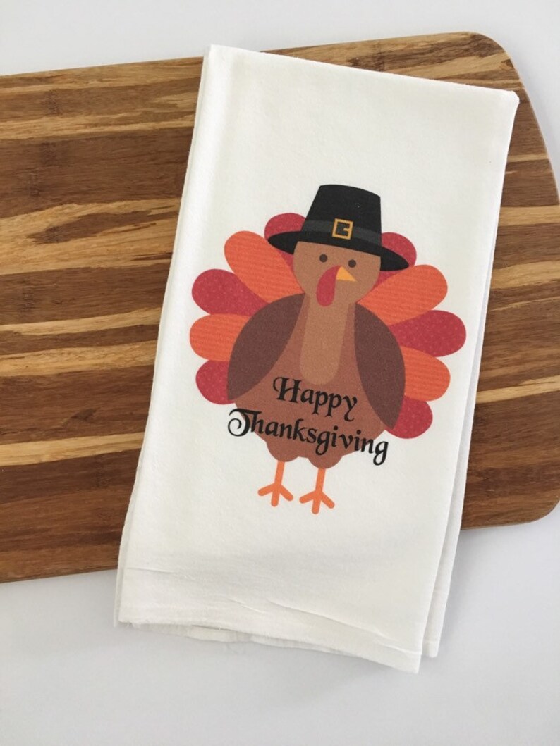 Puede incluir: Toalla de cocina blanca con un dibujo de un pavo de dibujos animados que lleva un sombrero de peregrino. El pavo tiene plumas marrones y una papada roja y naranja. El texto "Happy Thanksgiving" est&aacute; impreso debajo del pavo.