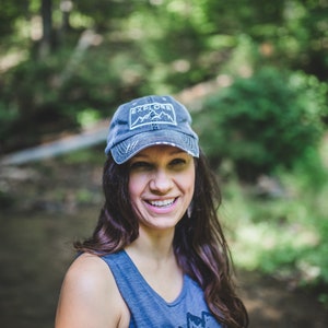 May include: A woman wearing a blue denim baseball cap with the word "EXPLORE" and a mountain graphic on the front. She is smiling and looking to the right.