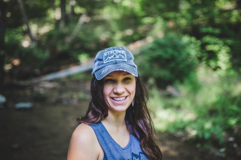 May include: A woman wearing a blue denim baseball cap with the word "EXPLORE" and a mountain graphic on the front. She is smiling and looking to the right.