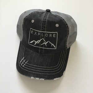 May include: A black and gray distressed trucker hat with a white embroidered mountain range and the word "EXPLORE" in a rectangle.