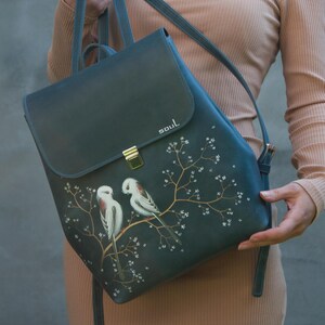 Könnte beinhalten: Ein dunkelblauer Lederrucksack mit handgemaltem Motiv von zwei weißen Vögeln auf einem blühenden Zweig. Die Klappe hat einen goldfarbenen Metallverschluss und das Wort "soul" in Weiß. Der Rucksack verfügt über verstellbare Schultergurte.