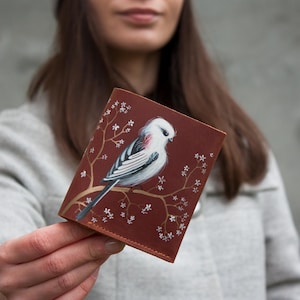 May include: A brown leather wallet featuring a hand-painted bird perched on a branch with white flowers. The bird has white, black, and red markings. The wallet is rectangular and appears to be closed.