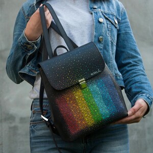 以下が含まれることがあります： レインボーデザインの黒いバックパック。フラップには「soul」の文字がプリントされています。レインボーはカラフルなドットで構成されています。バックパックにはシルバーの留め金と黒いストラップが付いています。