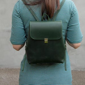 Könnte beinhalten: Ein dunkelgrüner Lederrucksack mit Klappenverschluss und goldfarbenem Verschluss. Der Rucksack hat verstellbare Träger und einen oberen Griff. Die Person auf dem Bild trägt ein grün-weiß gestreiftes Kleid.