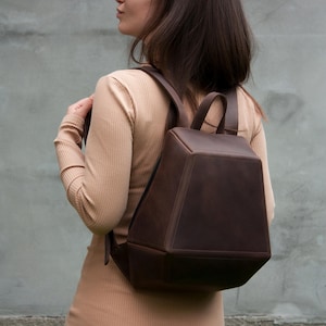 May include: A dark brown leather backpack with a structured, geometric, trapezoidal shape. The bag features a flat front panel, a top handle, and two adjustable shoulder straps. The surface has a matte finish with visible stitching along the edges.