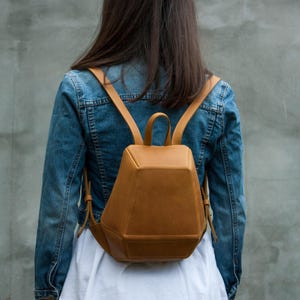 May include: A mustard yellow backpack with a structured, geometric, multi-faceted design. The bag features a top handle, two thin shoulder straps, and visible stitching along the edges, worn over a blue denim jacket and a white shirt.