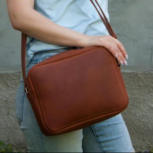 May include: A rectangular brown leather crossbody bag with rounded corners, featuring a smooth matte finish, a top zipper closure, and a thin matching leather shoulder strap. The bag has a clean, structured design with visible stitching along the edges.