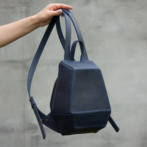 May include: A dark blue geometric backpack with a structured, multi-faceted shape. The bag features a top handle, adjustable shoulder straps, and a zippered closure. The design is composed of several angular panels joined by visible stitching.