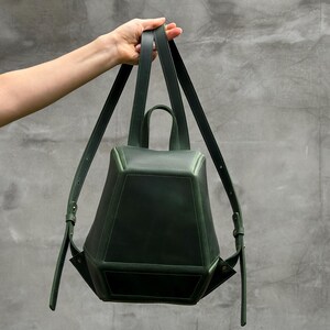 May include: A dark forest green backpack with a structured, geometric, multi-faceted shape. The bag features a top handle, two adjustable shoulder straps, and a smooth, matte finish. The design consists of angular panels joined together.