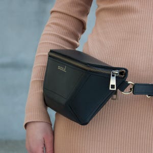 May include: Black fanny pack with a gold zipper and the word "SOUL" printed on the front. The bag is worn around the waist. The person is wearing a long-sleeved, ribbed, tan dress.