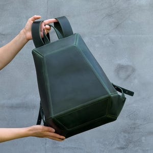 May include: A dark forest green backpack with a structured, geometric, multi-faceted design. The bag features a smooth matte finish, a top handle, and adjustable shoulder straps. The construction consists of angular panels joined by visible seams.