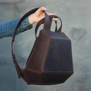May include: A geometric, dark brown leather backpack with a unique, angular design. Features a top handle, adjustable shoulder straps, and a zippered closure. The bag is held up by a person's hand.